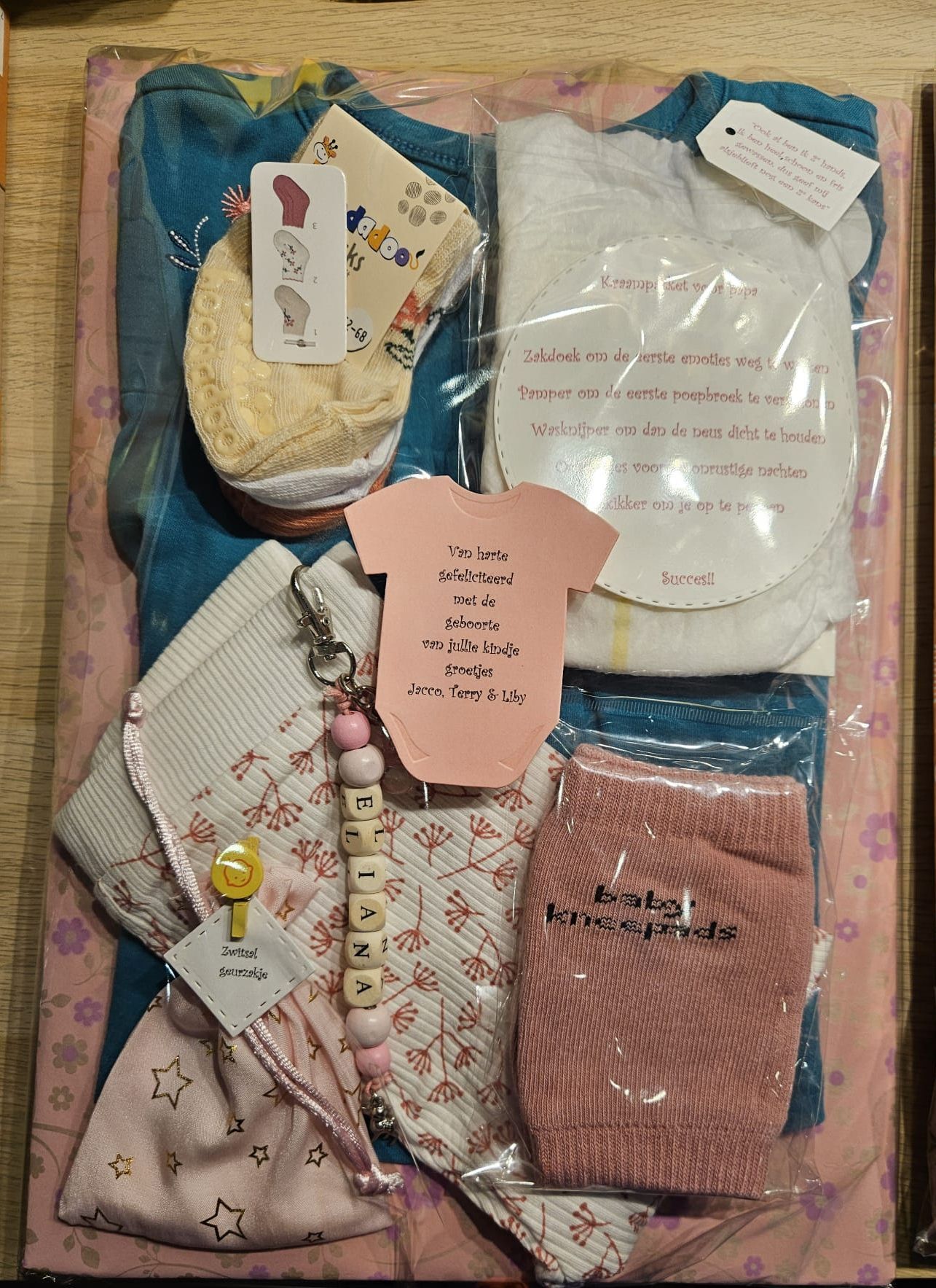 Vier opgevouwen babykleertjes in een geschenkdoos met een roze bedankkaartje, pastelkleurige en gestreepte stoffen.