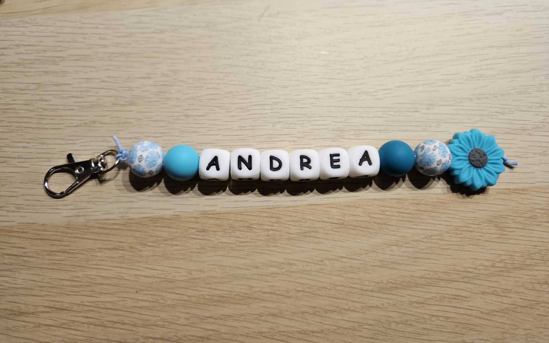 Blauwe kralen sleutelhanger met witte letterkralen die ANDREA spellen en een bloemhanger op een houten oppervlak.