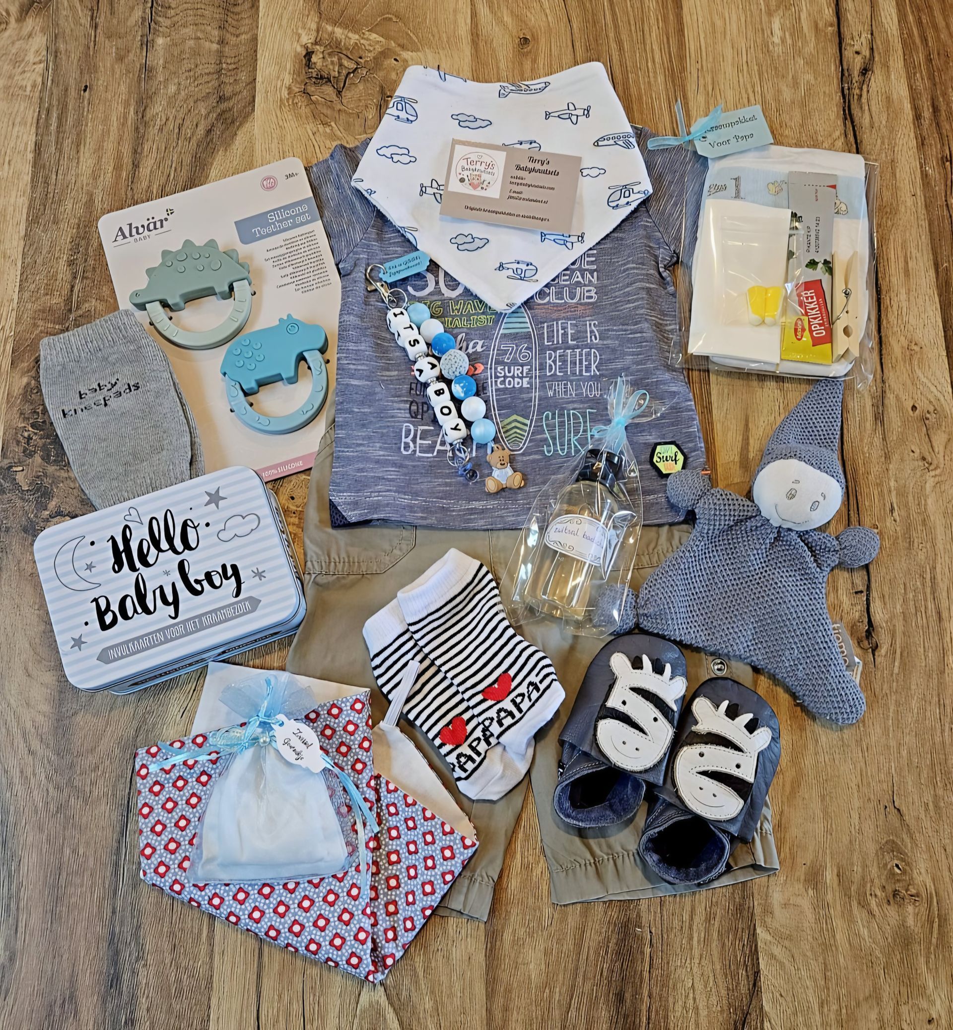 Een gebreide babyoutfit, bestaande uit een blauw vestje, babyschoentjes, slabbetjes, een mutsje en een speenhouder, ligt op een houten vloer.