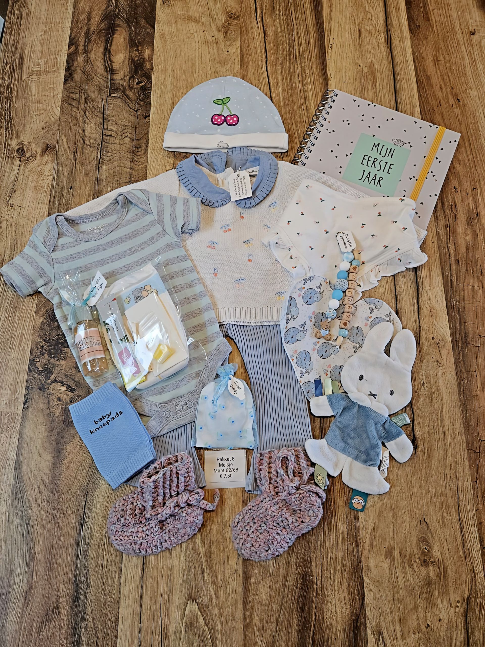 Babykleertjes en accessoires uitgestald op een houten vloer, waaronder rompertjes, mutsjes, sokjes, slabbetjes en babyschoentjes.