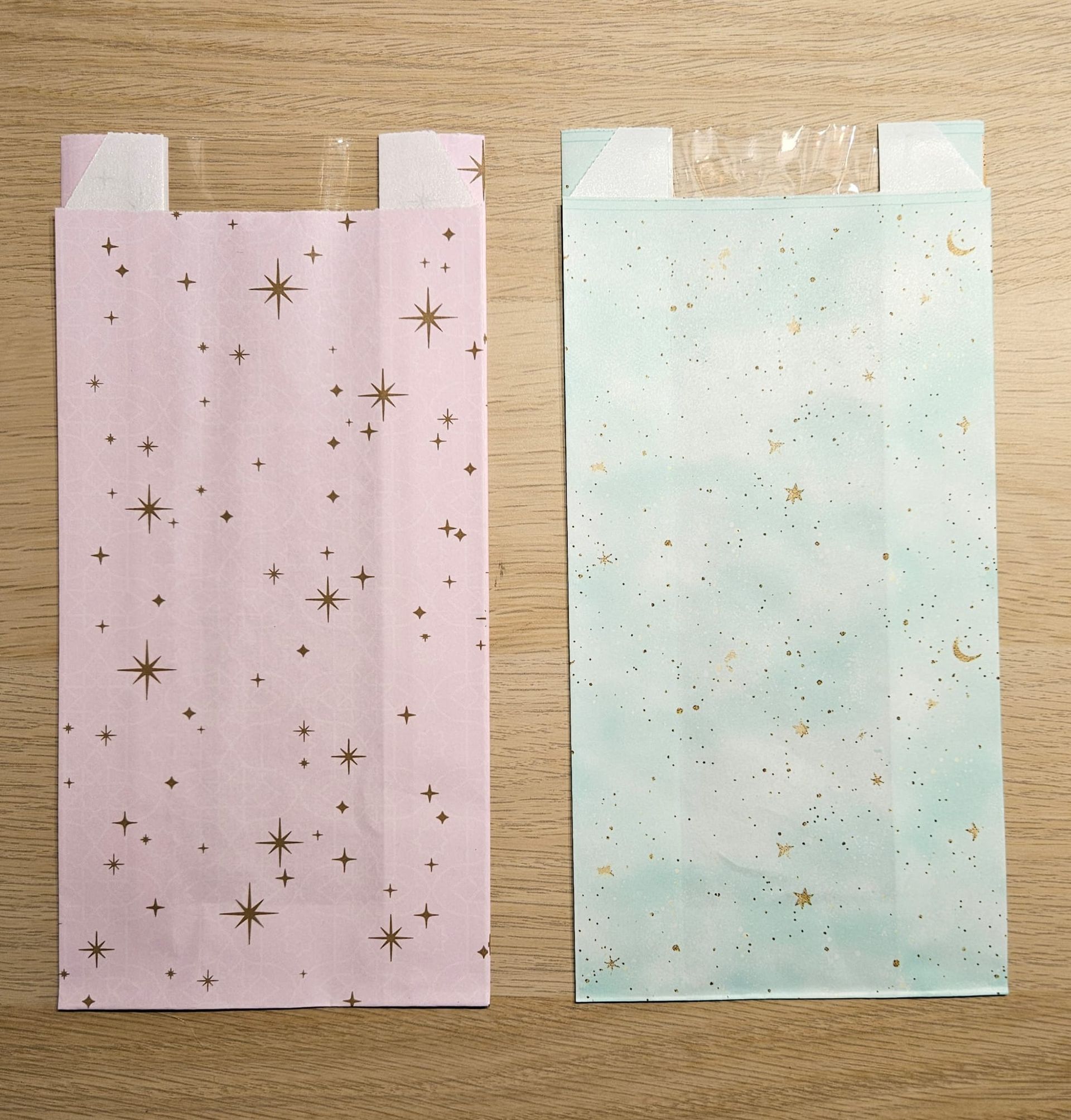 Twee pastelkleurige cadeauzakjes, roze met gouden sterren en mintgroen met gouden spikkels, op een houten ondergrond.