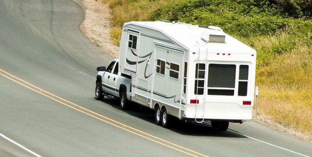 Un camion blanc tractant un camping-car blanc le long d'une route sinueuse à deux voies ; végétation verte et jaune sur le bord de la route.