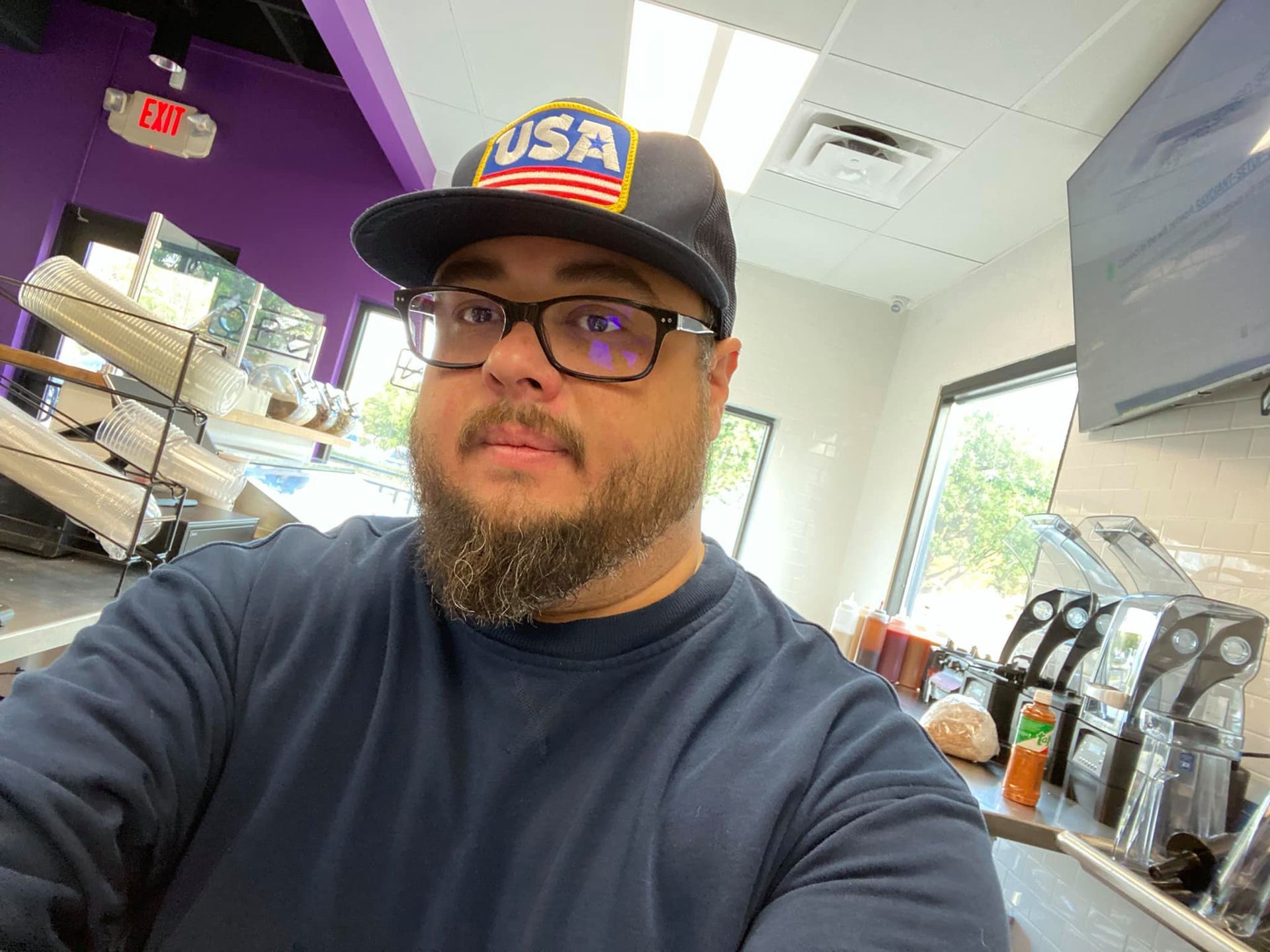 A man with a beard wearing a hat and glasses is taking a selfie in a kitchen.