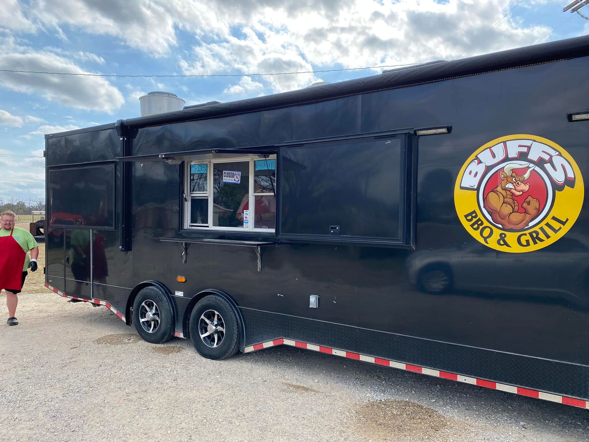 A black food truck with a yellow logo on it is parked in a gravel lot.