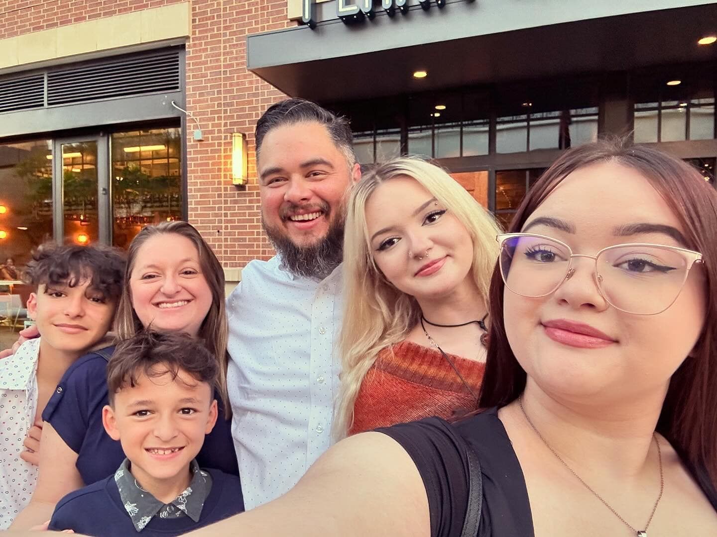 Group selfie of six smiling people standing outside a brick building at dusk