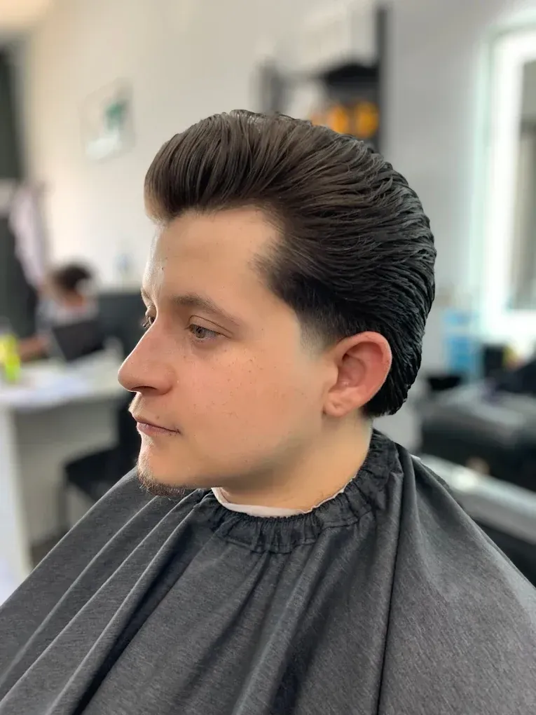 Man with a pompadour haircut, fade on the sides, wearing a barber cape in a salon.