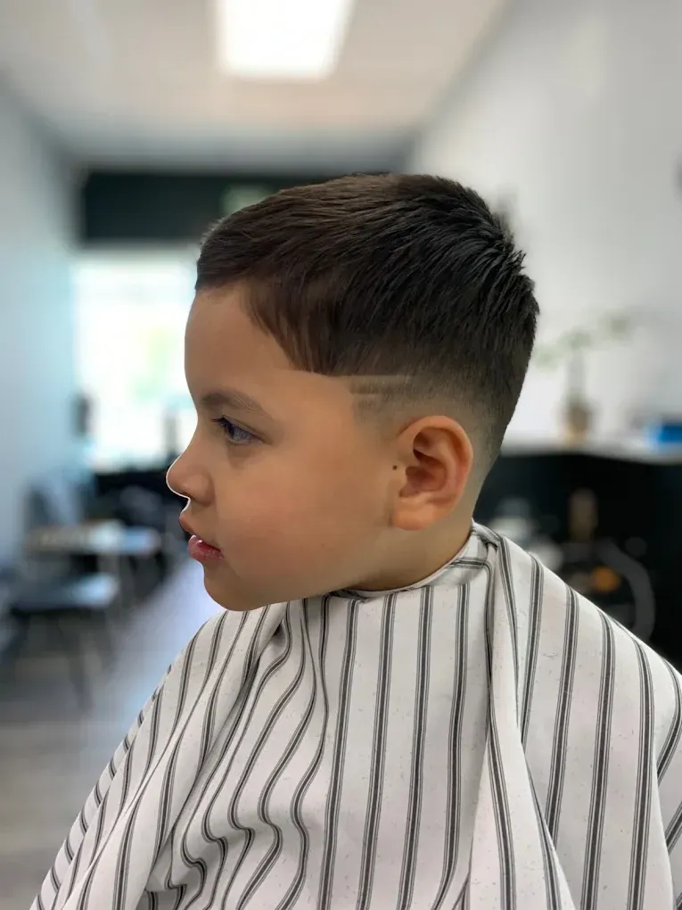 Young boy with a fresh haircut, fade with a line design, wearing a barber cape, in a salon.