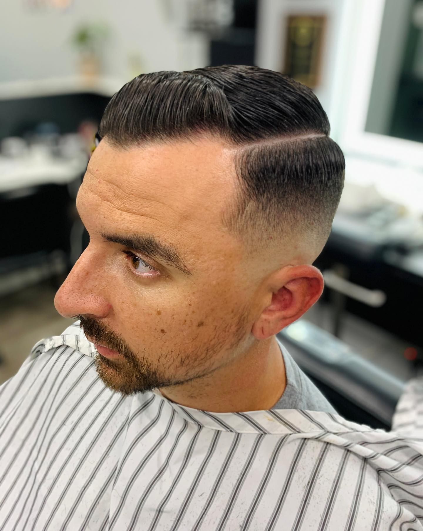 Man with a side part, short hair and a skin fade. He has a neatly trimmed beard and is wearing a barber cape.