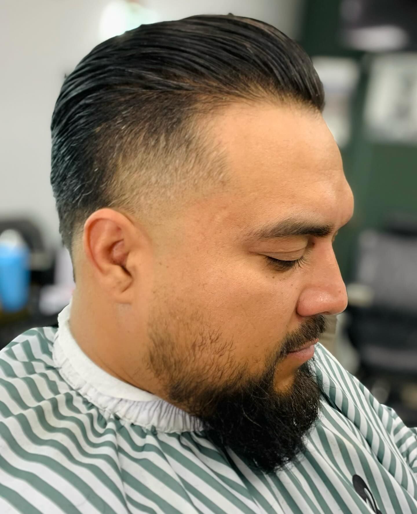 Man with slicked-back hair and faded sides, trimmed beard, wearing a striped barber cape.
