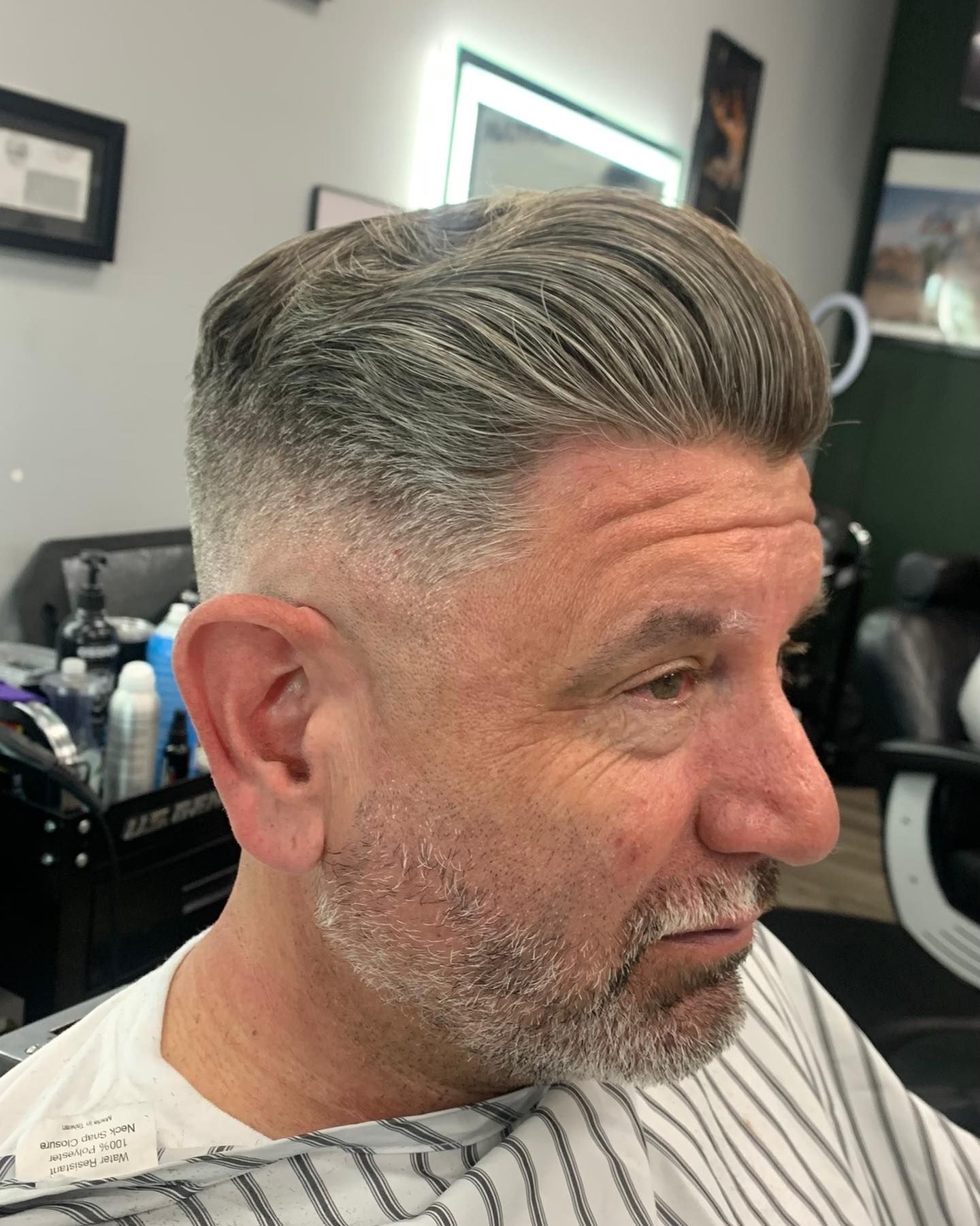 Man with a faded haircut, styled back. Gray hair, stubble, wearing a barber's cape.