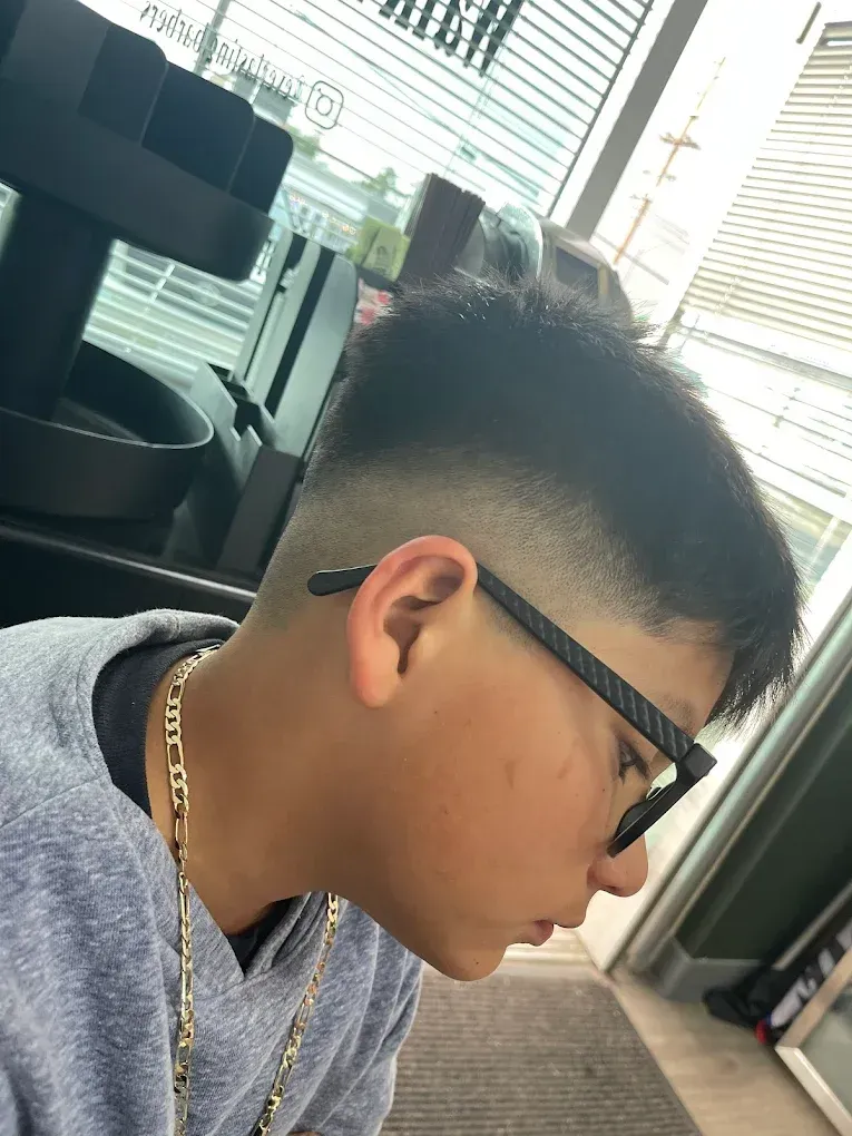 Person with a fade haircut and glasses in a building, wearing a sweater and chain necklace, looking down.