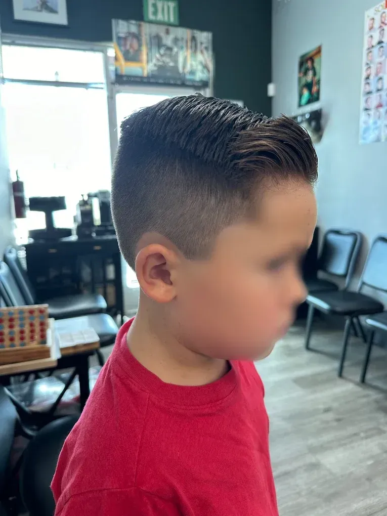 Boy with dark hair in a fade haircut, in a barber shop setting. He is wearing a red shirt.