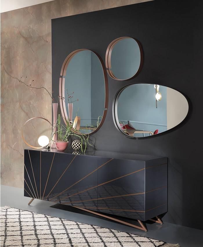 Mobiletto nero con dettagli in rame, tre specchi ovali e lampada decorativa contro la parete scura.
