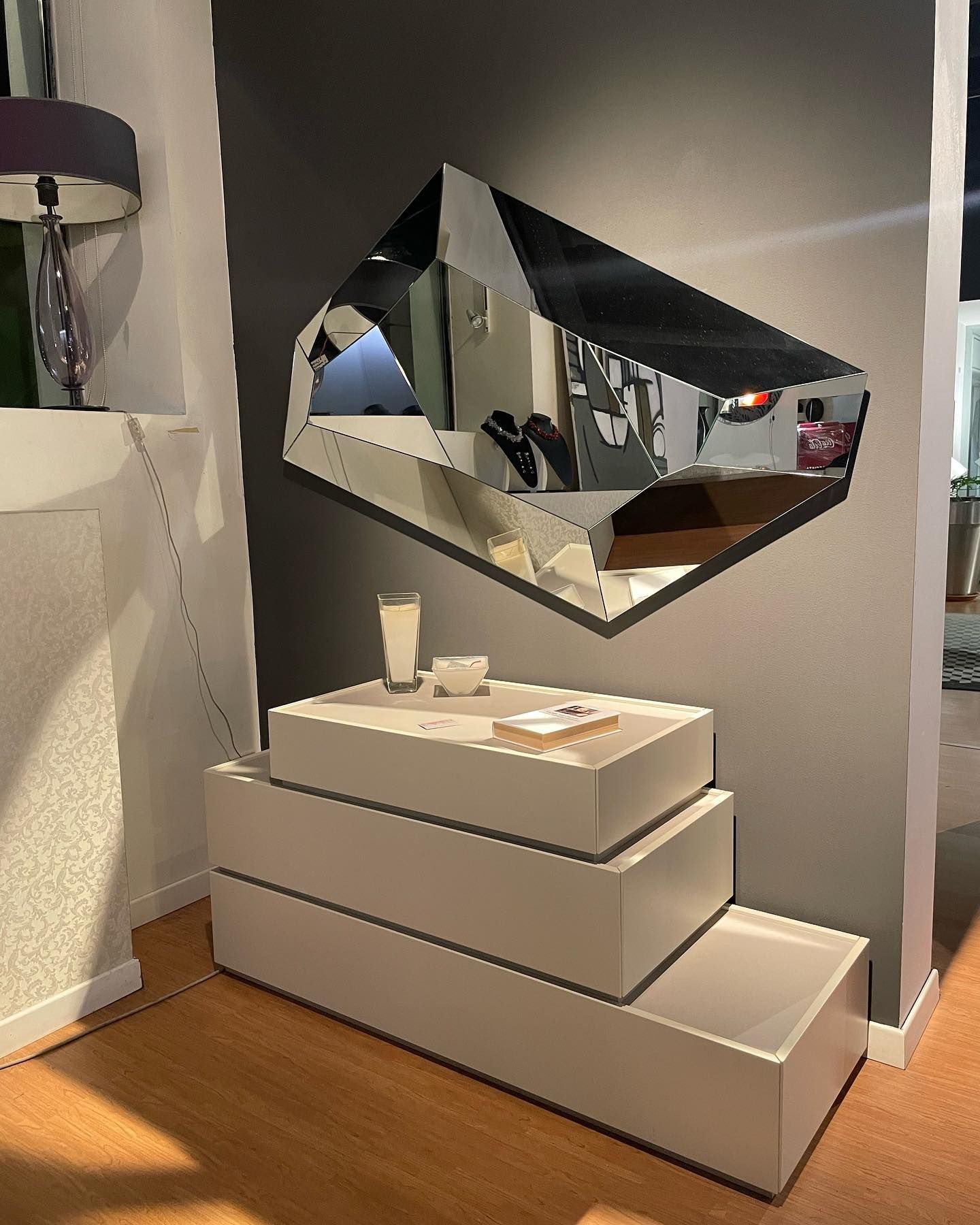 Specchio geometrico sopra un mobile contenitore bianco a più livelli. Parete grigia, lampada e pavimento in legno sullo sfondo.