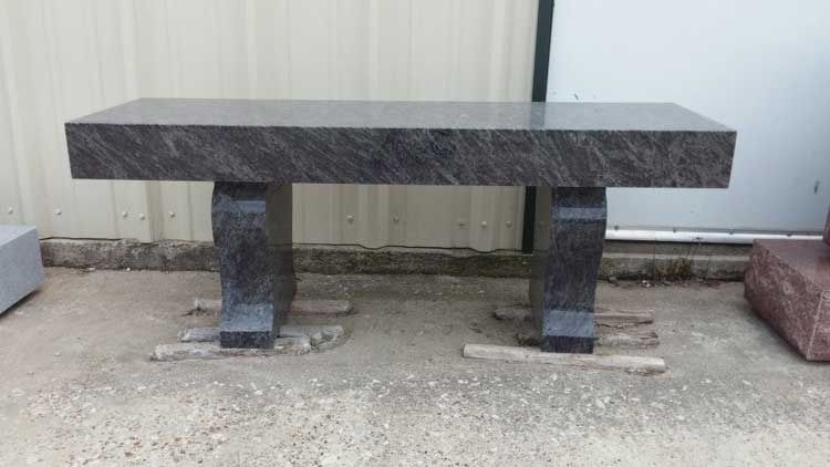 Dark stone bench with two block legs on gravel beside a corrugated metal wall