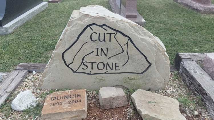 Stone sign engraved “CUT IN STONE” in a cemetery, with smaller memorial stones at its base