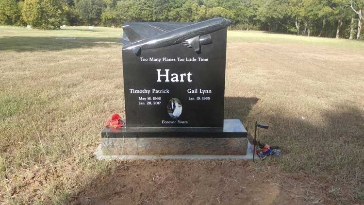 Black memorial gravestone in a grassy field with flowers and a small flag, surrounded by trees.