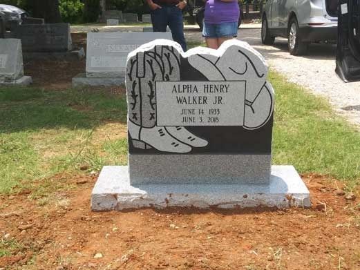 Gravestone with engraved tree design in a grassy cemetery, with a car and people in the background.