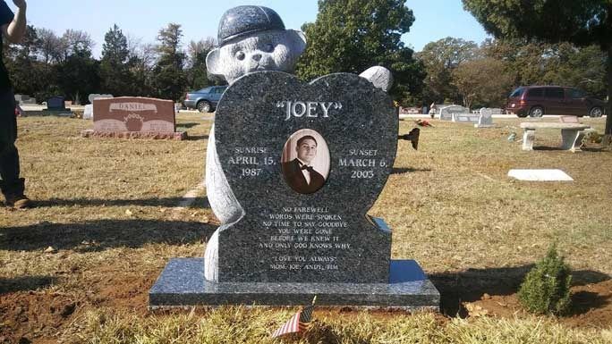 Gravestone with the name “JOEY” and a portrait photo in a cemetery, with a large statue behind it.