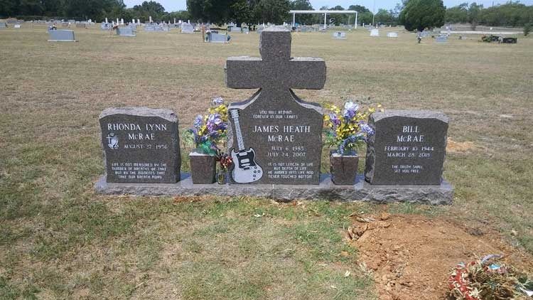 Three grave markers with flowers in a cemetery, centered on a large cross-shaped headstone.