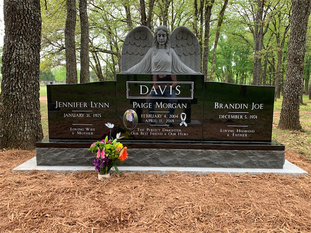 Black granite memorial headstone with angel carving and flowers in a wooded cemetery