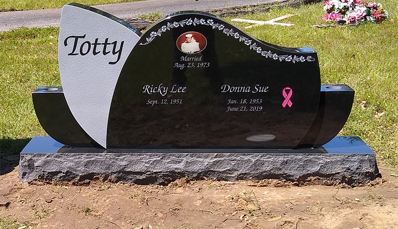 Black polished gravestone with pink ribbon and flowers, set on a stone base in a cemetery, engraved “Totty.”
