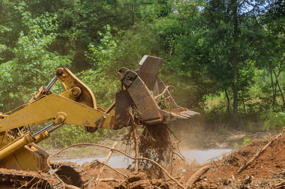 Yellow excavator clearing brush and roots in a muddy area, trees in the background.