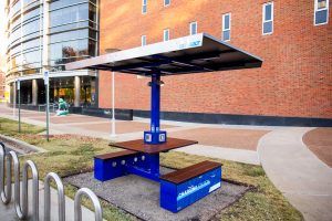A blue picnic table with a solar panel on top of it in front of a brick building.