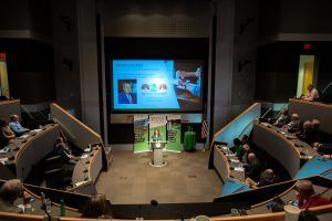 A group of people are sitting in an auditorium watching a presentation on a large screen.