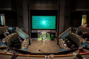 A group of people are sitting in a council chamber watching a presentation on a large screen.