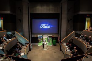 A ford logo is displayed on a large screen in a conference room.