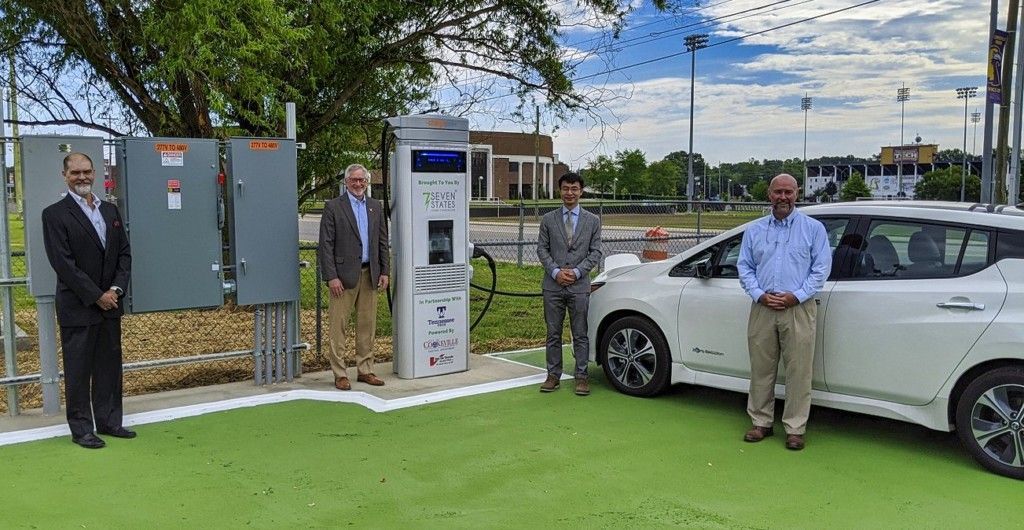 Three men are standing next to a white electric car at a charging station.