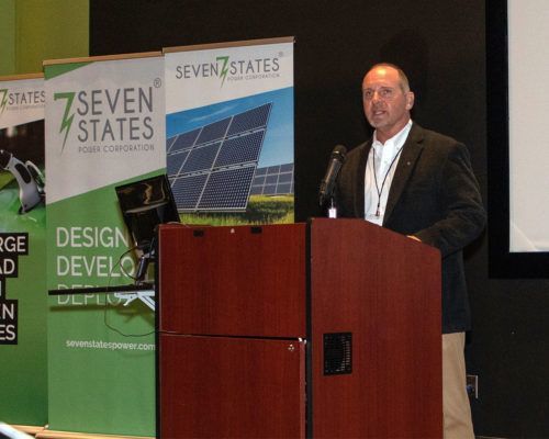 A man stands at a podium with seven states banners behind him