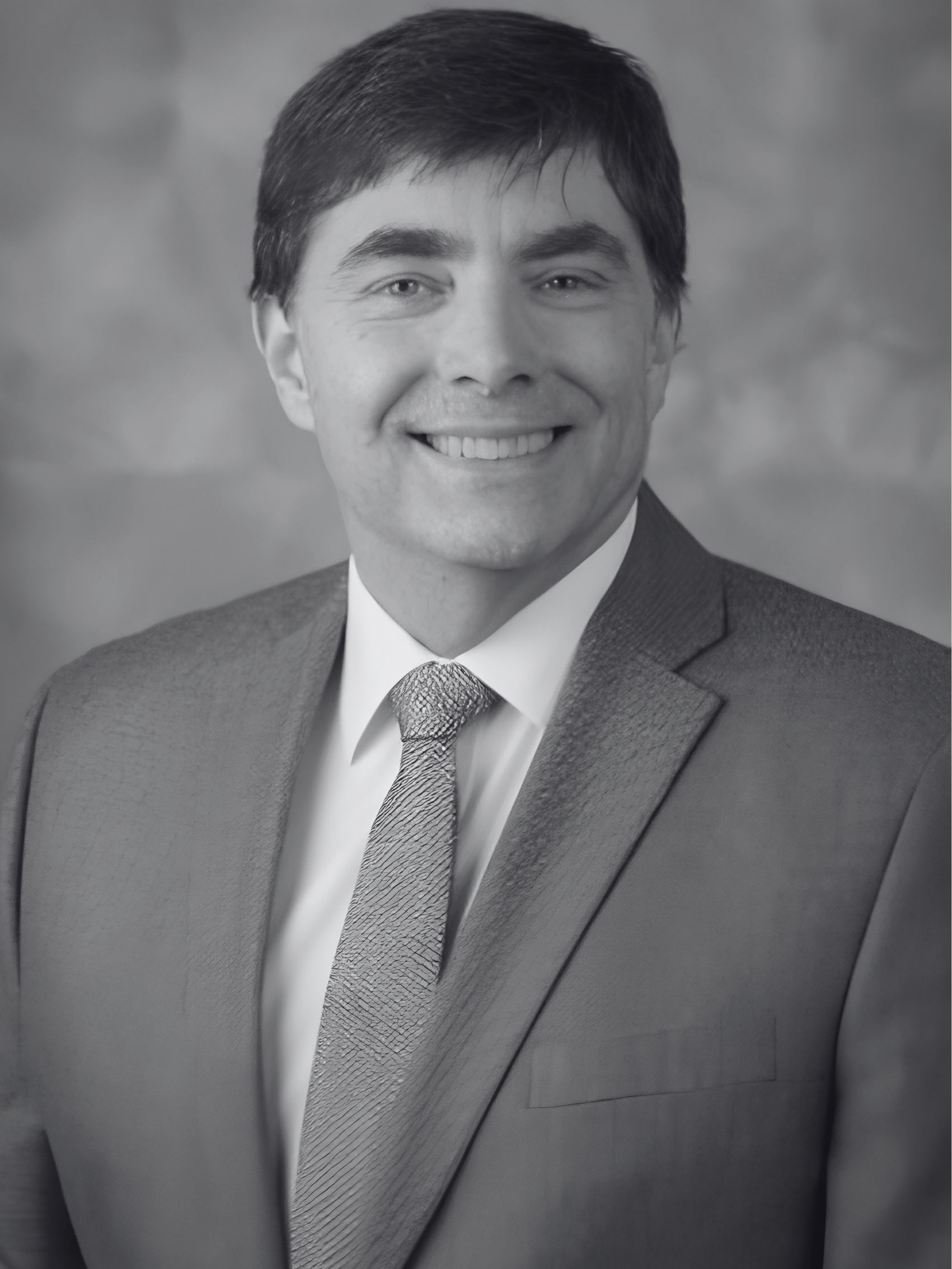 A bald man wearing glasses and a suit and tie is smiling in a black and white photo.