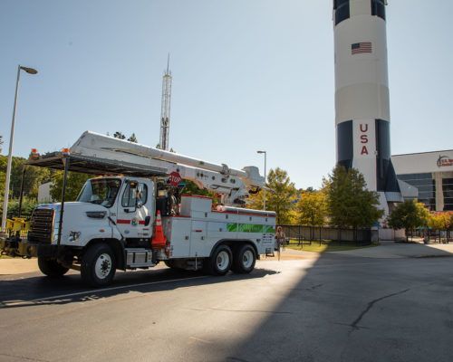 A white truck with a crane on top of it is parked in front of a rocket.
