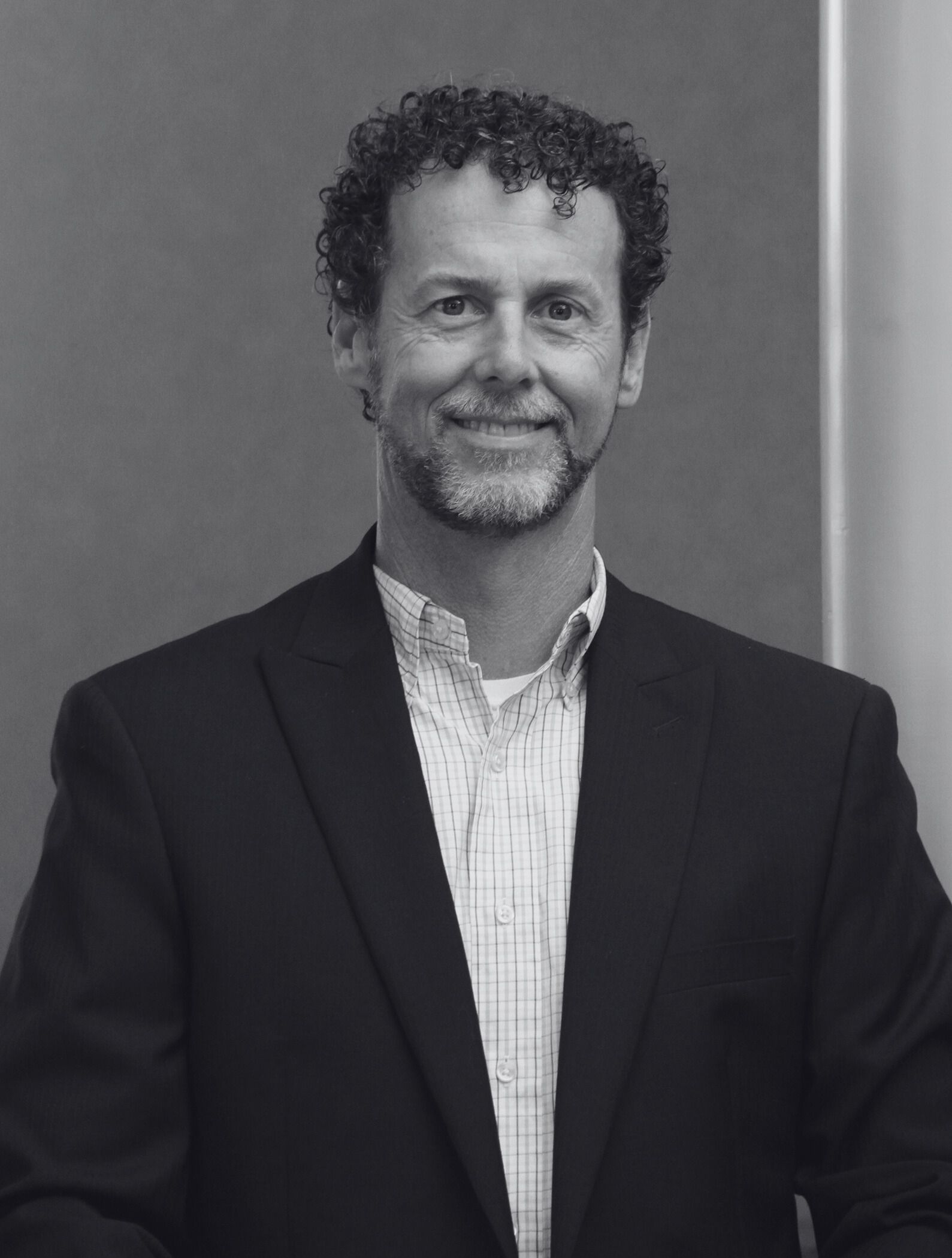 A man with curly hair and a beard is wearing a suit and smiling in a black and white photo.