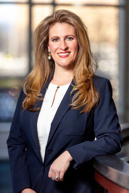 A woman in a blue jacket and white shirt is standing in front of a window.