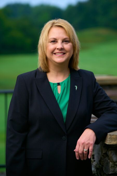 A woman in a black suit and green shirt is standing next to a stone wall.
