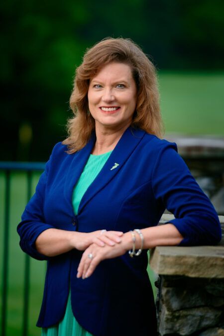 A woman in a blue jacket and green dress is leaning against a stone wall.