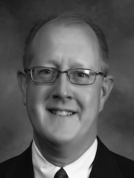 A man wearing glasses and a suit and tie is smiling in a black and white photo.