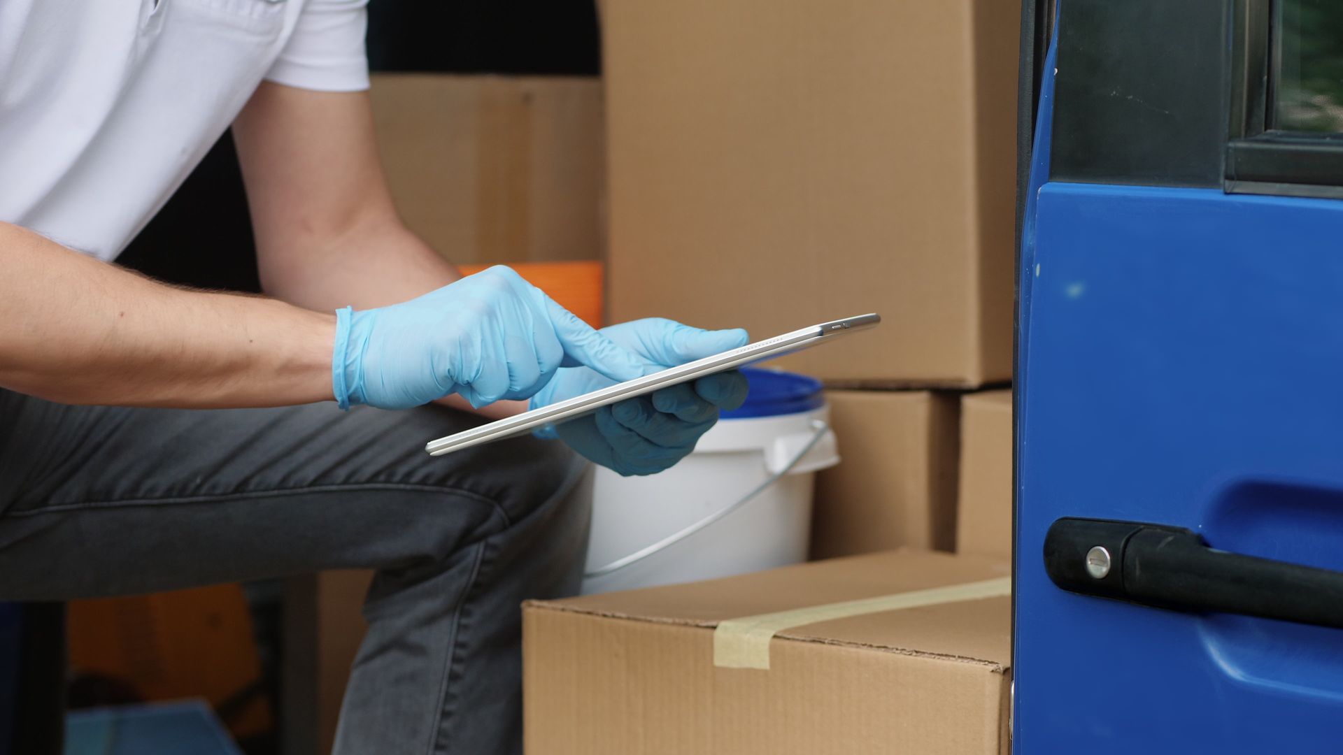 Medical mover in blue gloves using a tablet beside stacked cardboard boxes.
