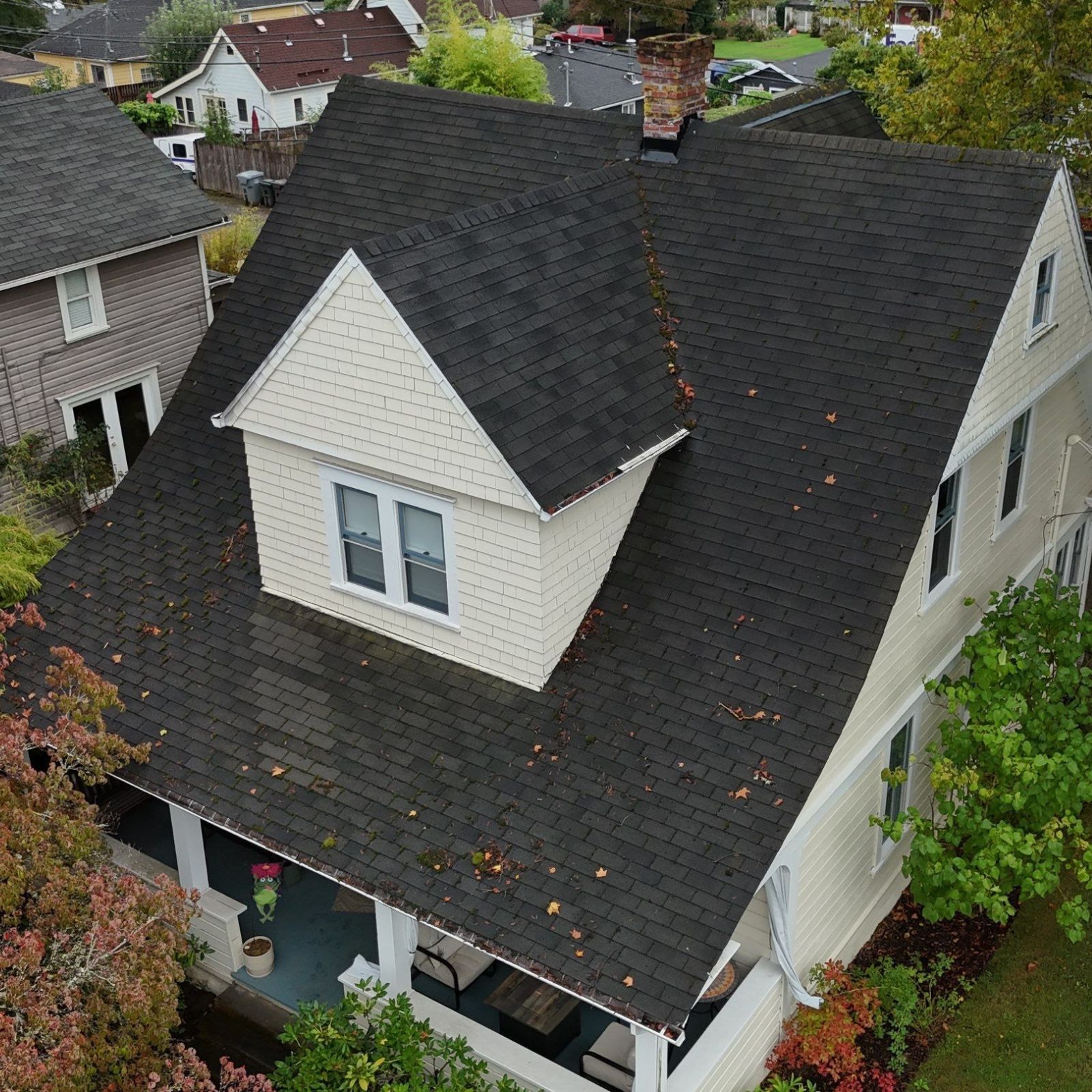 An aerial view of a two-story house with beige siding, a dark shingled roof, and a front porch, surrounded by green trees.