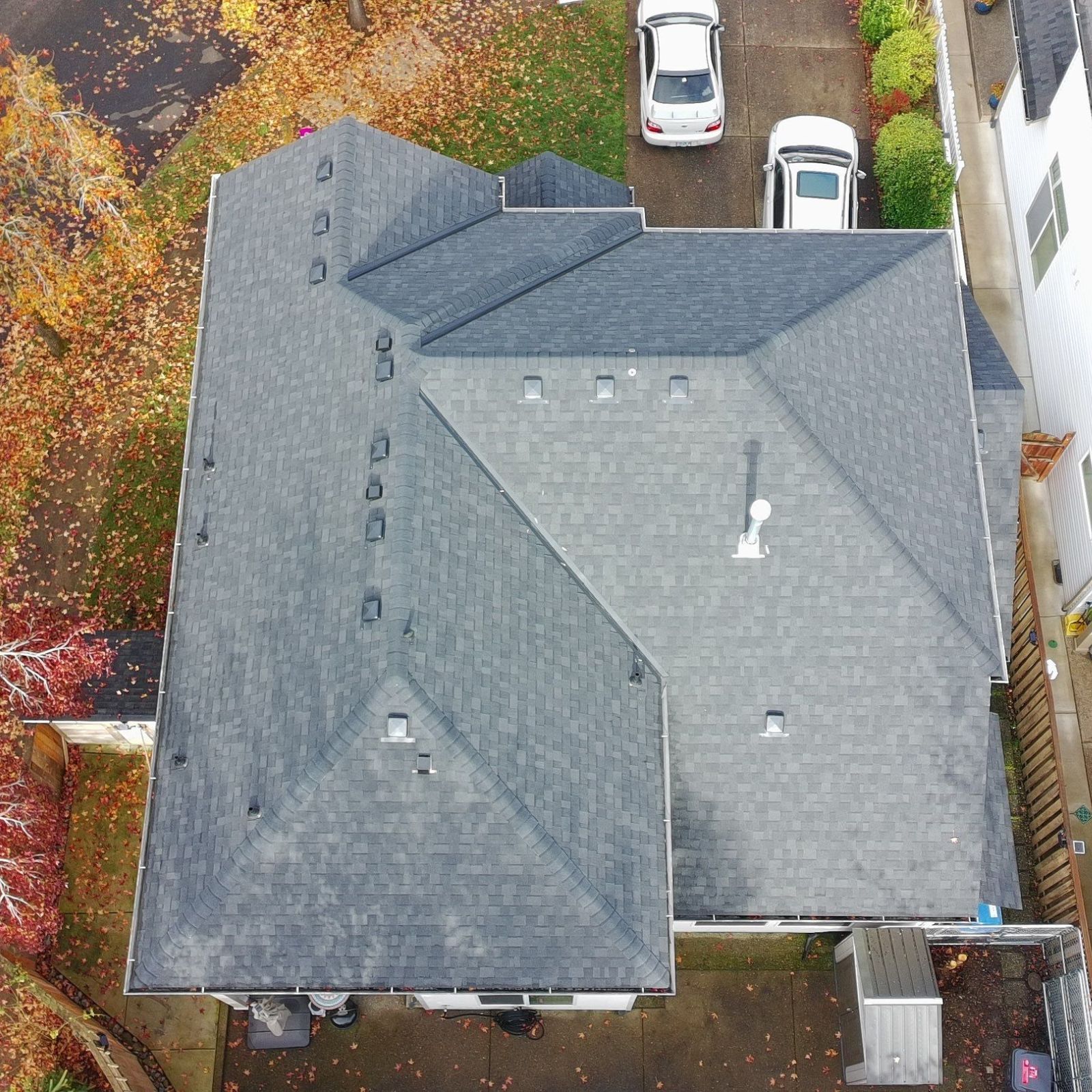 Aerial view of a gray shingled roof beside parked cars, surrounded by autumn trees.