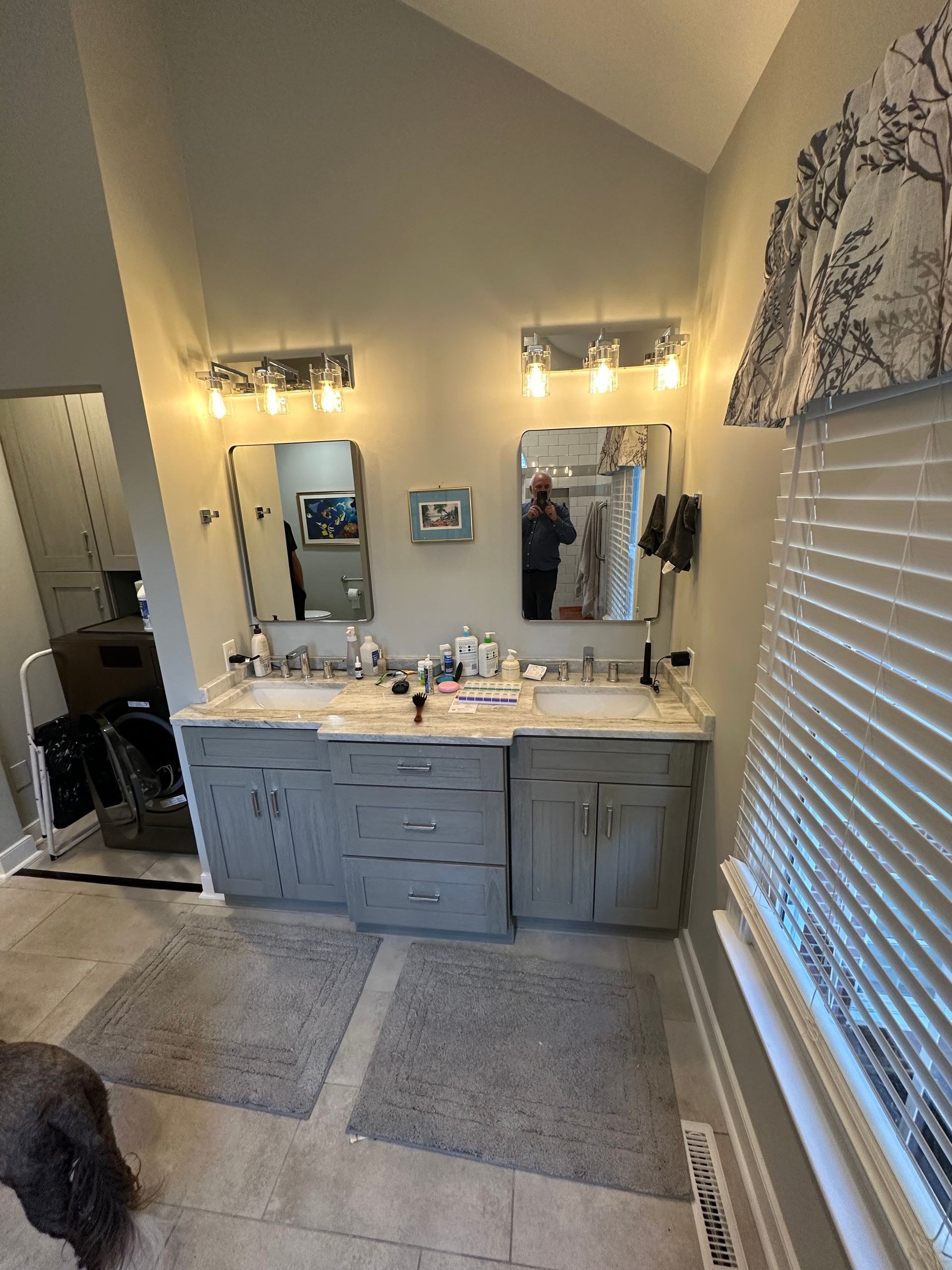 Bathroom with two sinks, gray cabinets, mirrors, and light fixtures. Blinds and a small window are on the right.