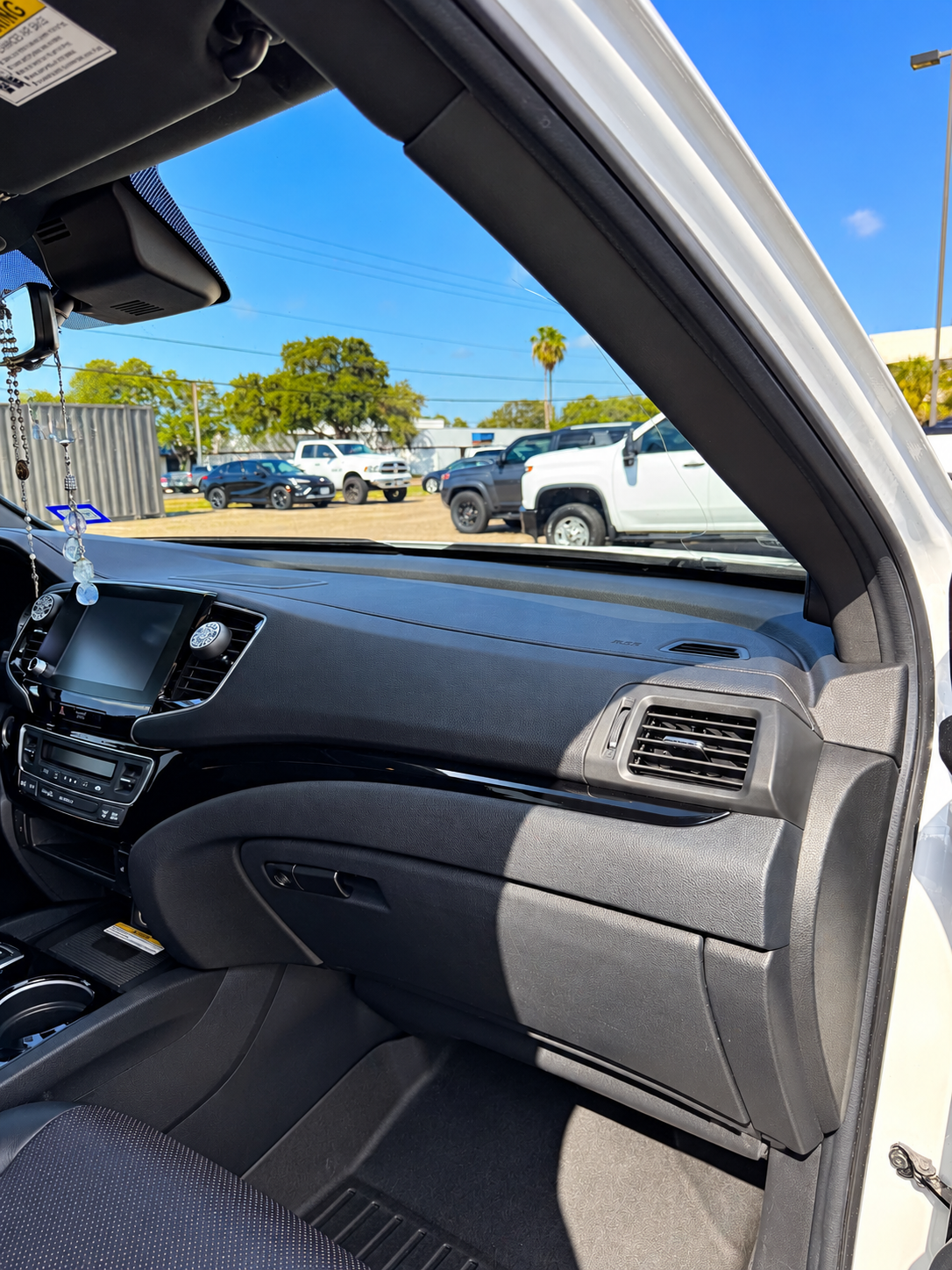 Car interior with dashboard and passenger door open to a sunny parking lot with trucks and palm trees.