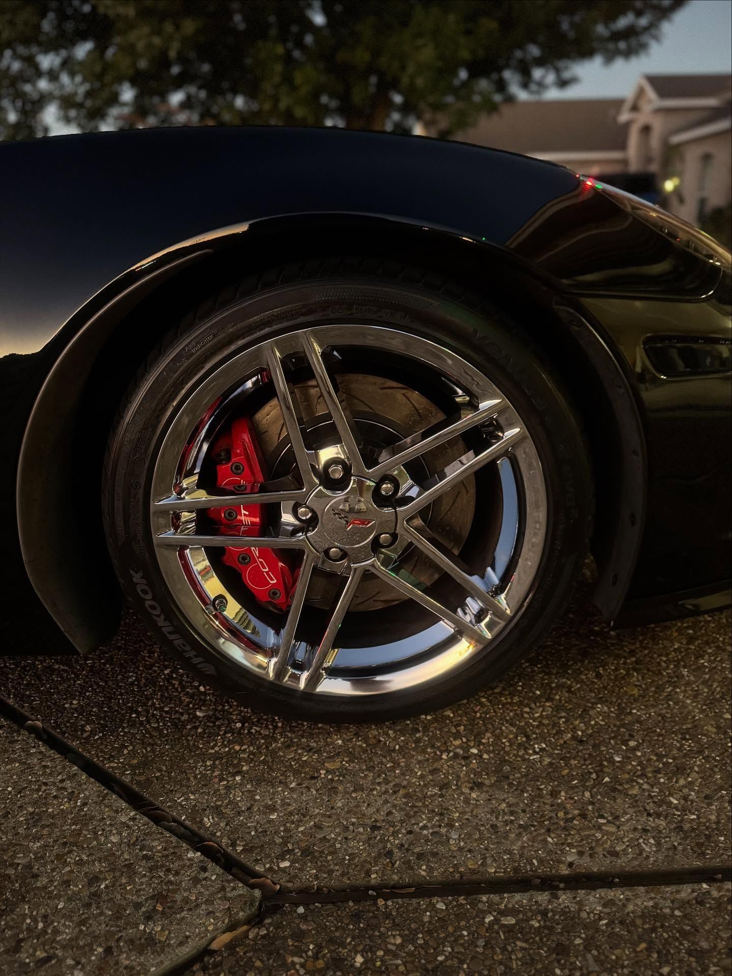 Close-up of a black sports car wheel with chrome rims and red brake calipers on wet pavement.