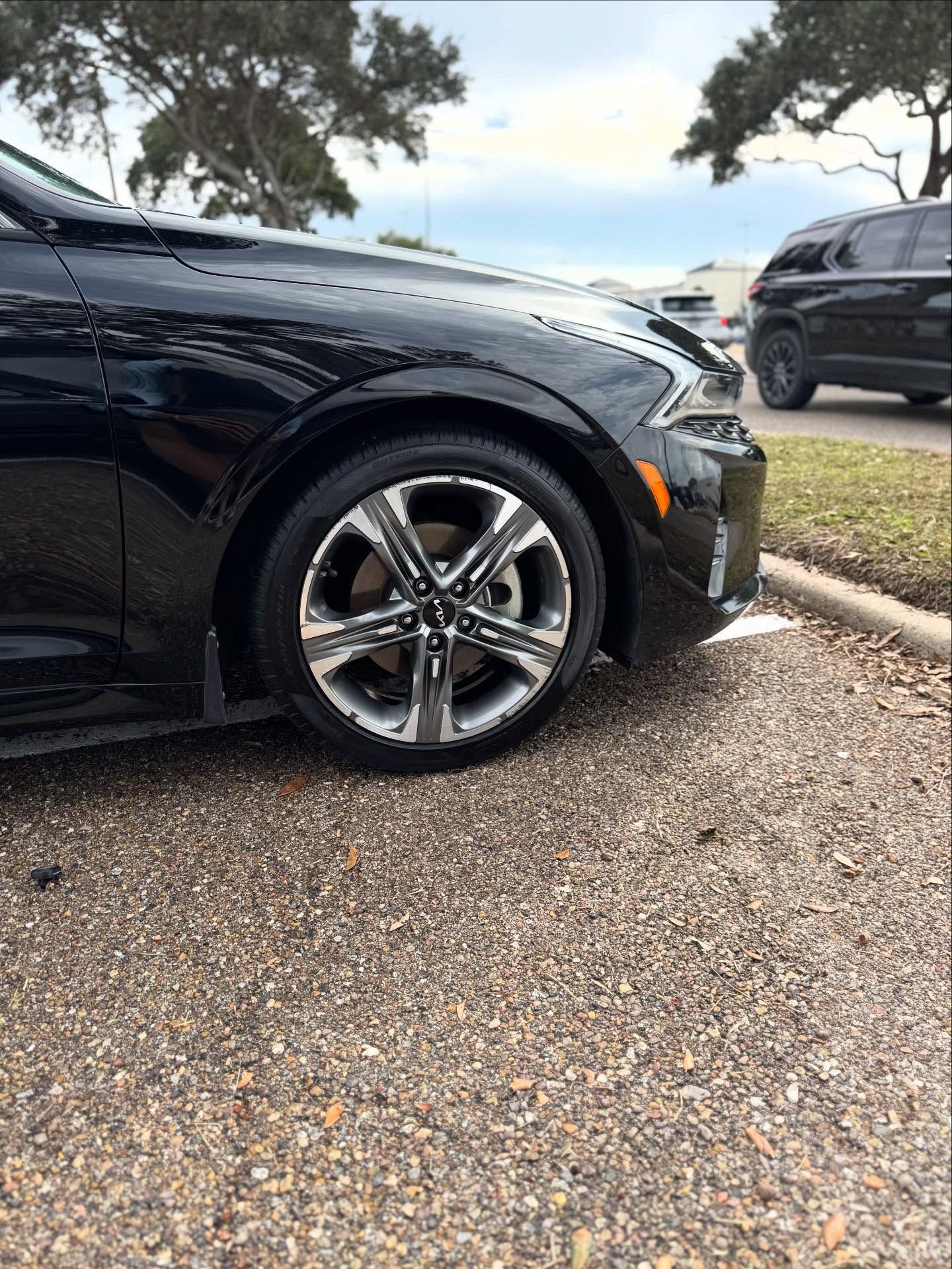 Black sedan parked on gravel, front wheel visible, with another dark SUV in the background.