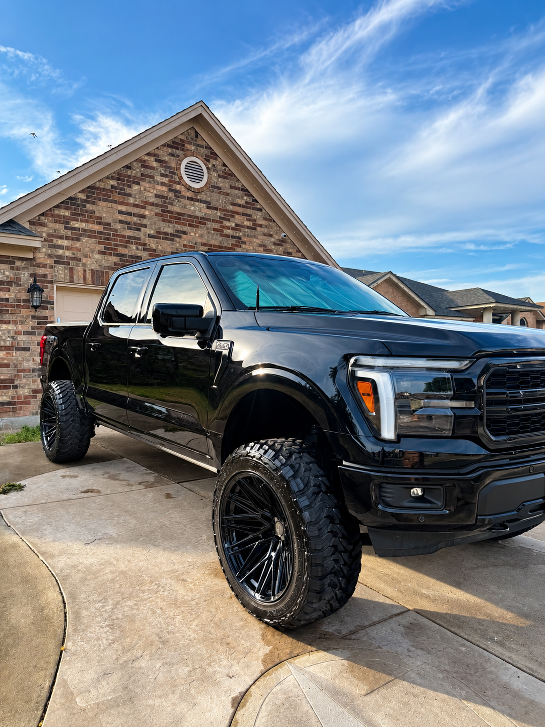 Black lifted pickup truck parked in a driveway beside a brick house under a blue sky
