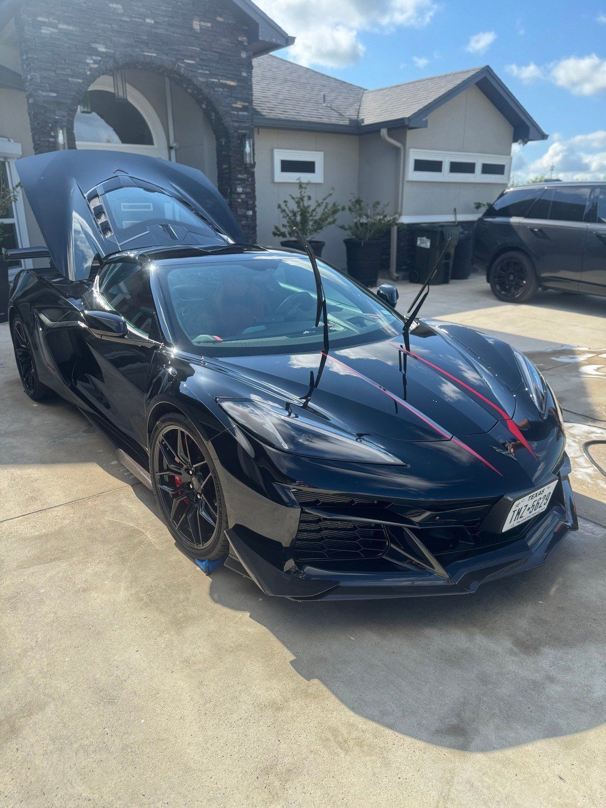 Black sports car with red accents parked in a driveway in front of a house with open hood and doors