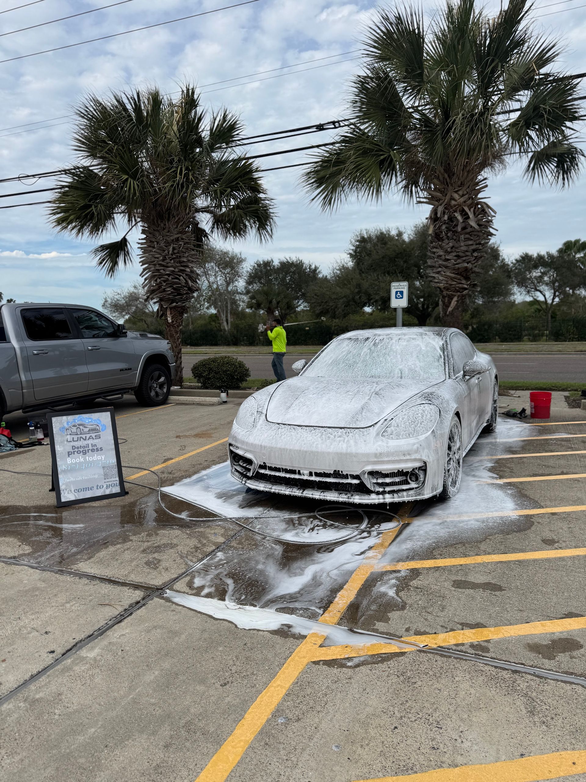 White car covered in foam at a car wash, with a person in a neon vest nearby and palm trees in the background